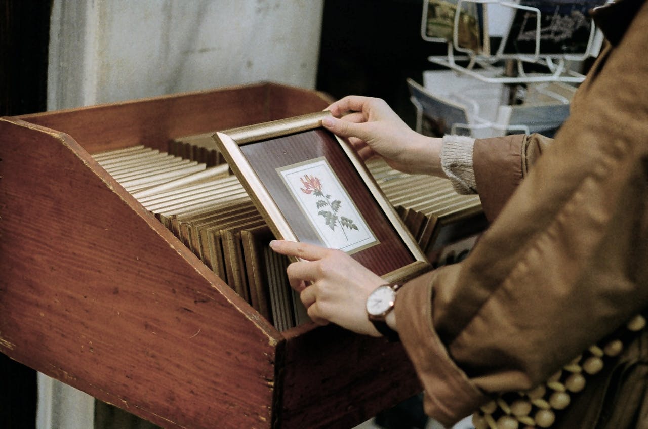 Side view of crop unrecognizable female shopper picking photo frame with flower above wooden box in old shop